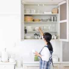 A woman wearing a blue plaid apron organizing a kitchen cupboard.