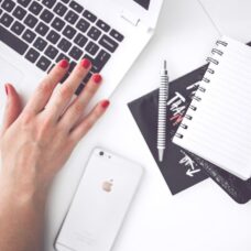 A woman's hand typing on a laptop with a cell phone, notepad, and pen on the desk.
