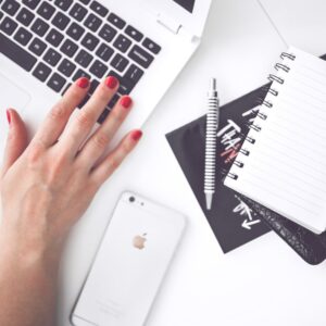 A woman's hand typing on a laptop with a cell phone, notepad, and pen on the desk.