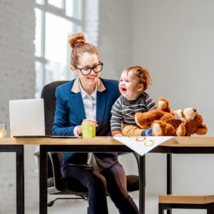 A busy mom sitting at a table with a laptop and her baby holding a teddy bear sitting on the table beside her smiling