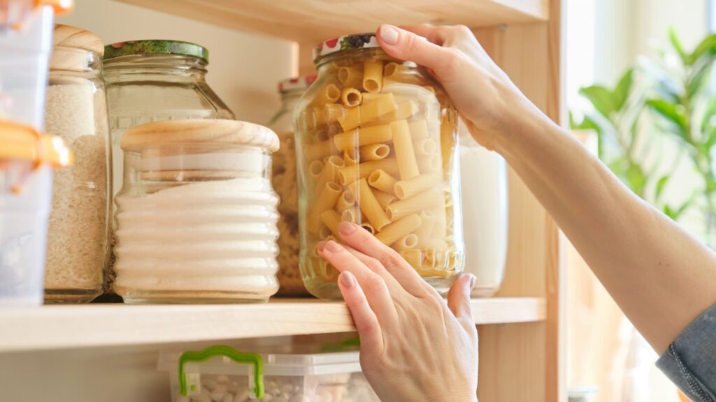 A woman's hands taking a clear jar of dried pasta off of a pantry shelf